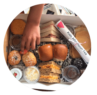 child's hand reaches into afternoon tea take away box to take a strawberry. Sandwiches, pancakes, scones and chocolates.