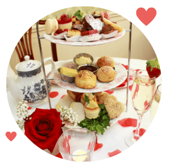 Valentines Day Afternoon Tea Stand with plates of petit fours, macarons, heart shaped chocolate brownies, buttermilk scones, sandwiches. Tea pot, glasses of prosecco and strawberries with antique chair in background. 
